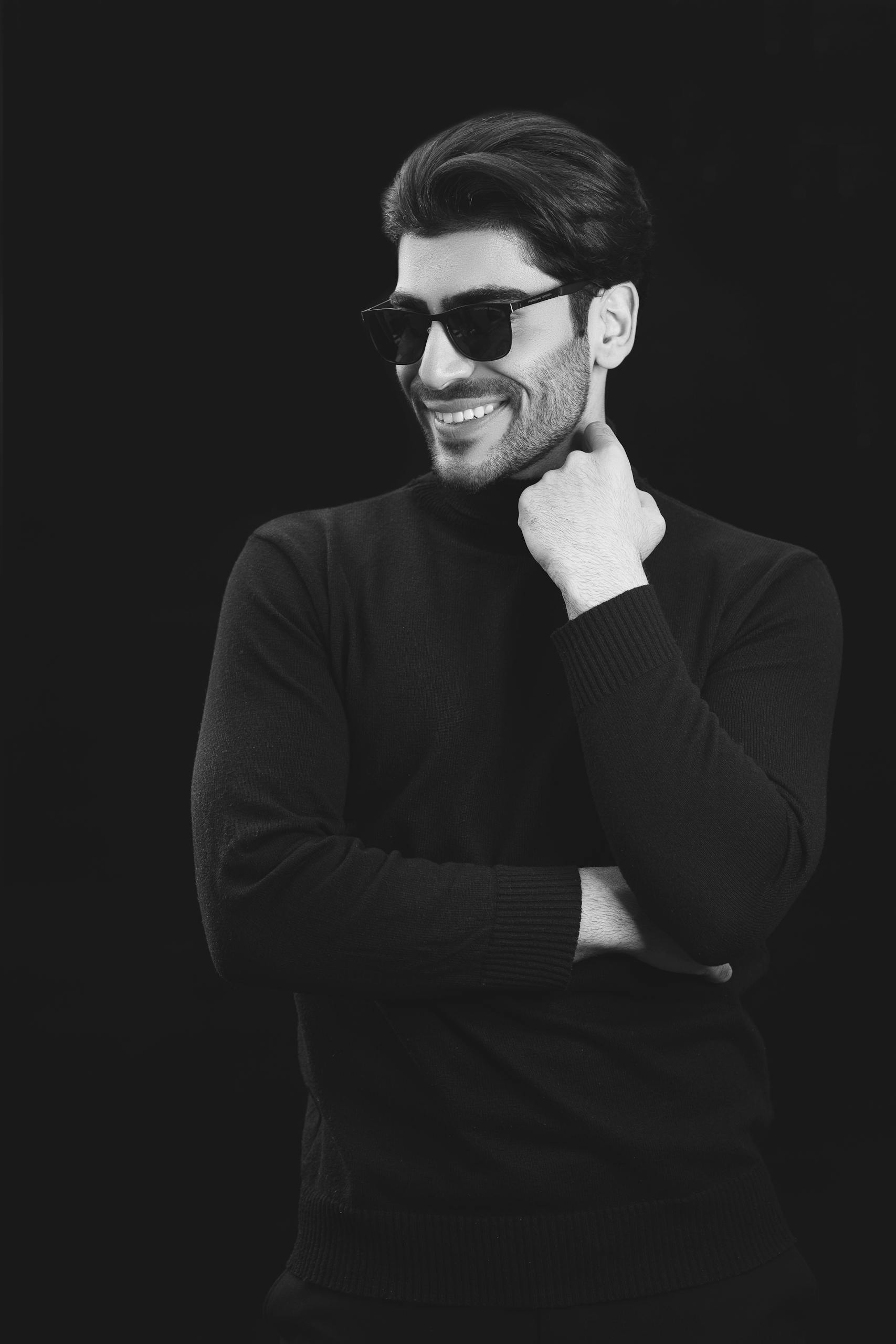 Monochrome portrait of a stylish man wearing sunglasses and a sweater, smiling confidently.